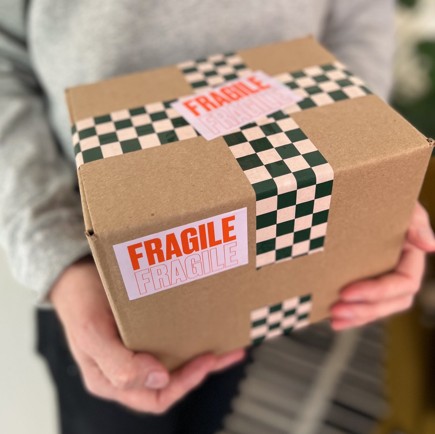 A person holding a box with checkered packing tape and pink fragile stickers.