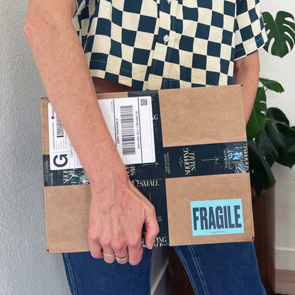 A woman in a checkered shirt holding a moving box with fragile sticker.