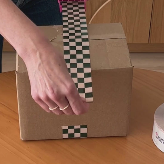 A woman is applying a strong and durable checkerboard pattern packing tape to a box.